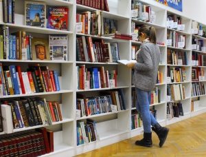 A woman in a gray sweater and blue jeans reading a book in front of a large white bookcase filled with books.