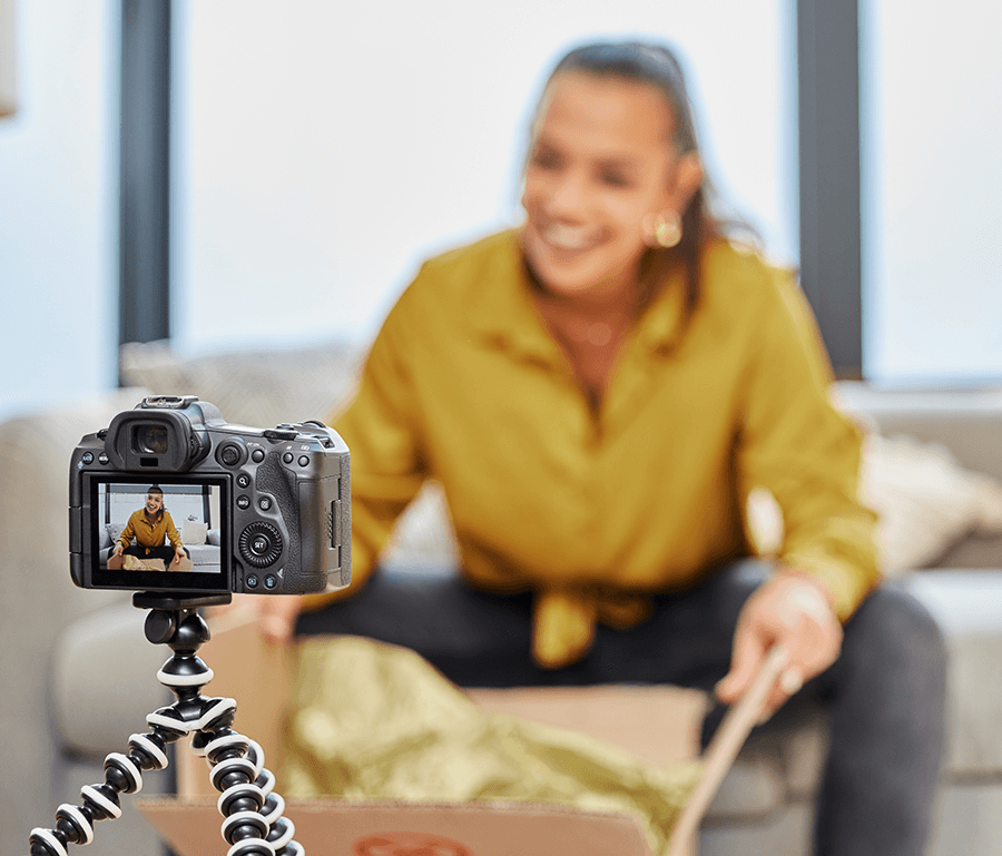 A woman sitting on s couch wearing a yellow blouse opening a box while being filmed. The video camera is the only thing focused while the rest of the image is blurred