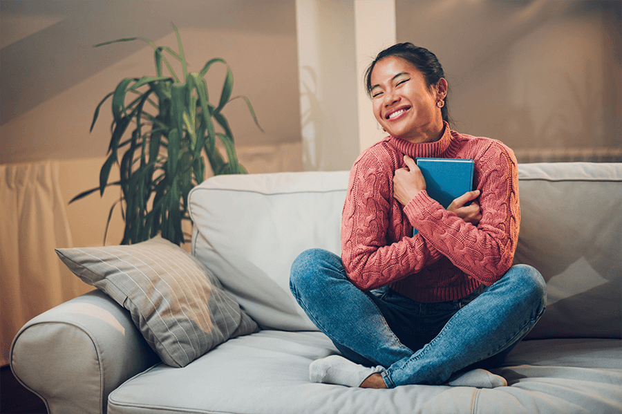 Young adult woman wearing a pink sweater sitting on her couch hugging a book close to her chest