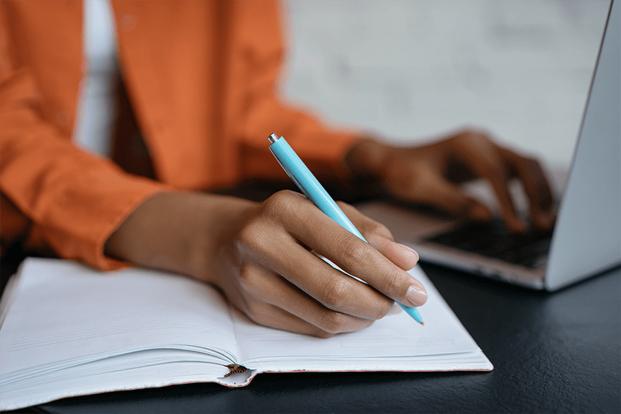 a closeup image of a woman's hand writing in a notebook using a bright blue pen. Her other hand is resting on top of her laptop.