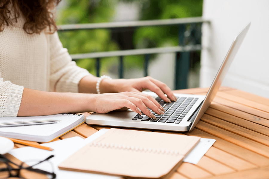 A woman with brown curly hair, wearing a white sweater is typing on a laptop with an open notebook next to her