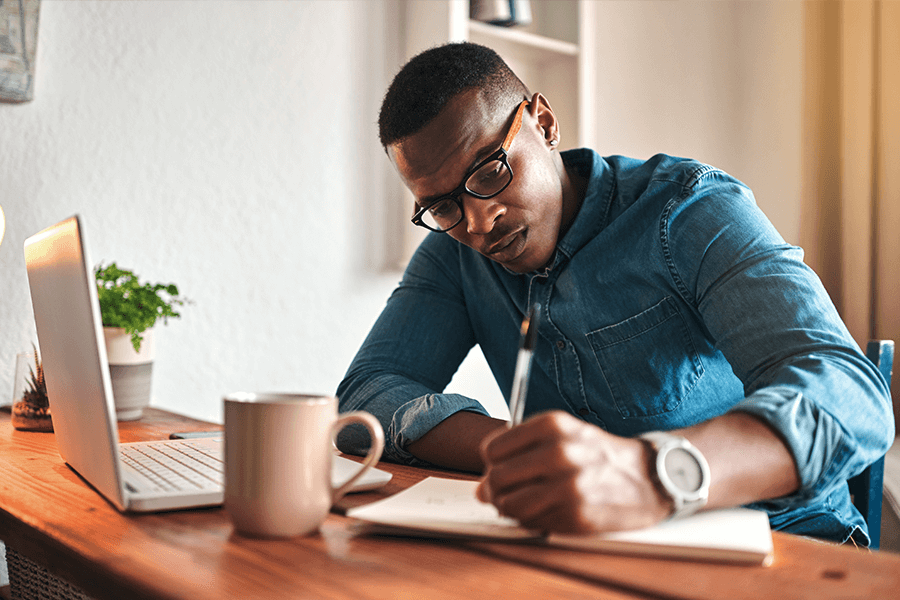 male author wearing a blue button up shirt, writing in a notebook next to his opened laptop and cup of coffee