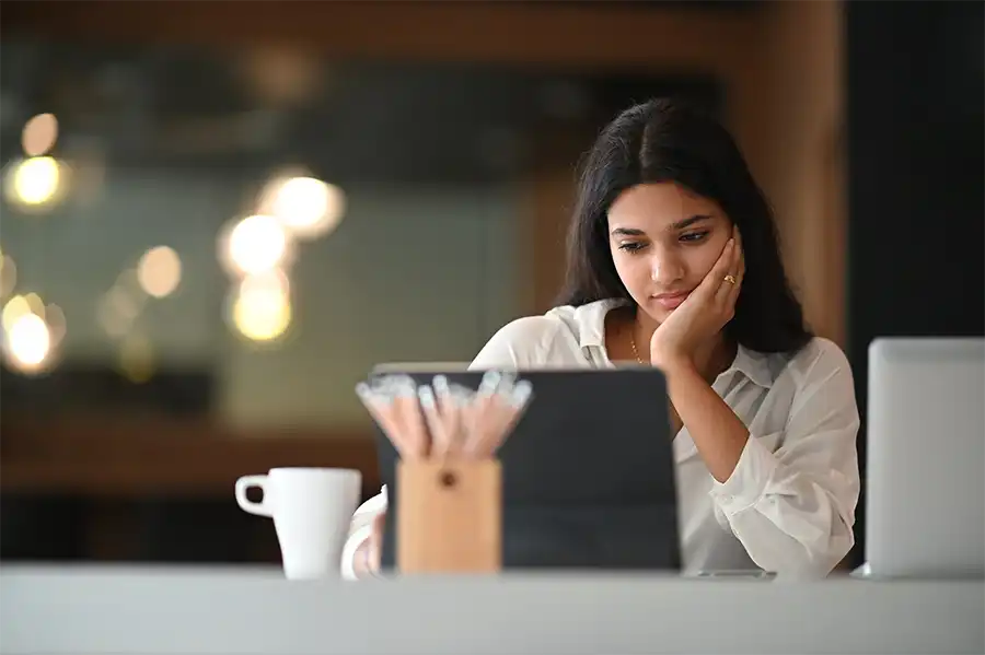 A young woman sitting at a desk looking at a tablet. Her facial expression shows she's a little sad.
