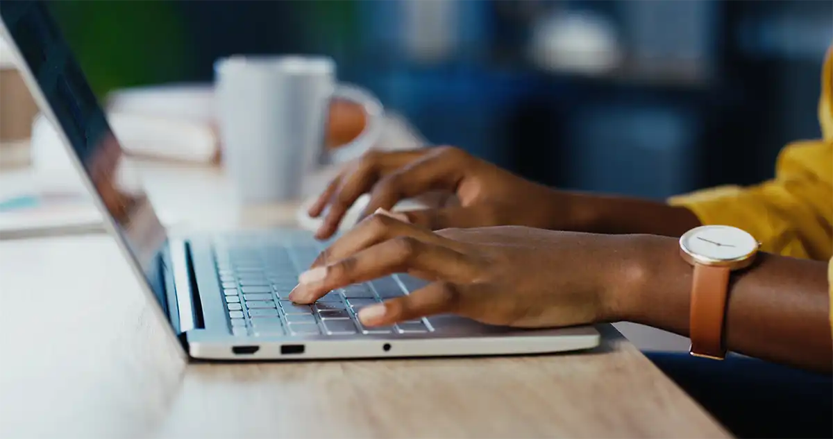 close up side view of a woman's hands typing on a laptop, the woman is wearing a yellow sweater and has a tan watch around her left wrist