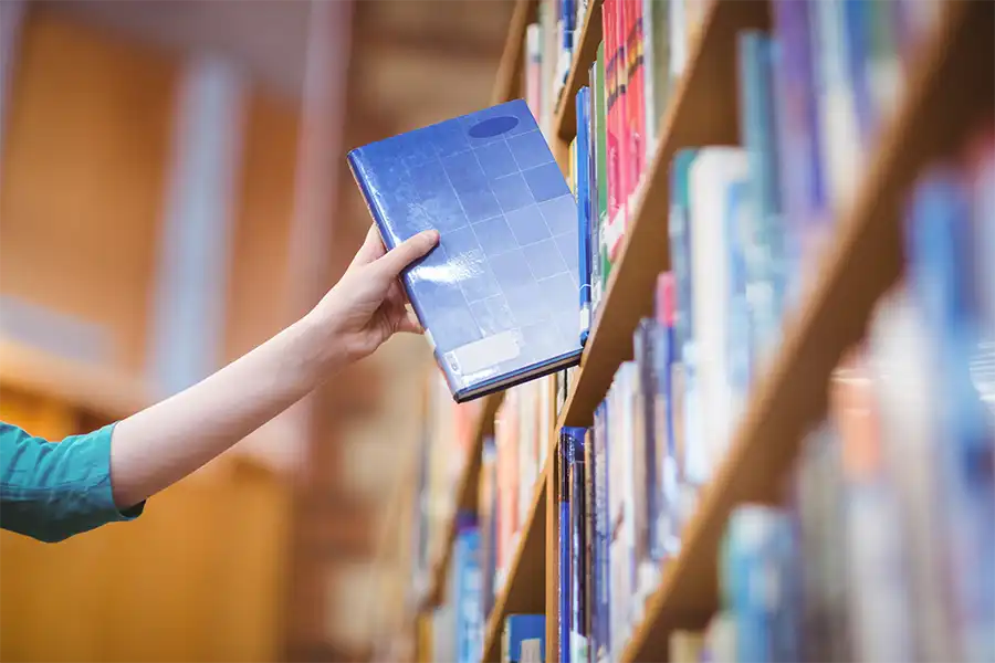 a young girl pulling a blue hard cover book out from a bookshelf in a library.