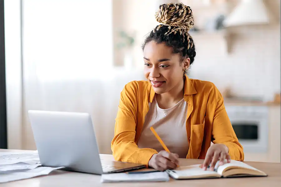 a young woman in an orange button up reading her laptop screen and taking notes in a notebook