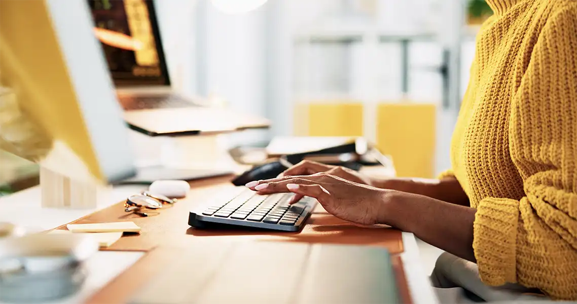 close up of a woman's hand typing on her bright yellow desktop computer, she is wearing a big sweater the same shade of yellow. Her desk, that her computer is. set up on, is made of light colored wood.