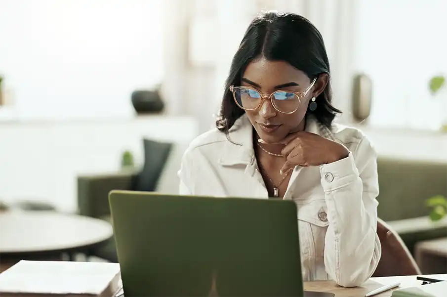 A young woman with black long hair and circle framed glasses is sitting at a light wood desk. She is working on a laptop with a green protective cover