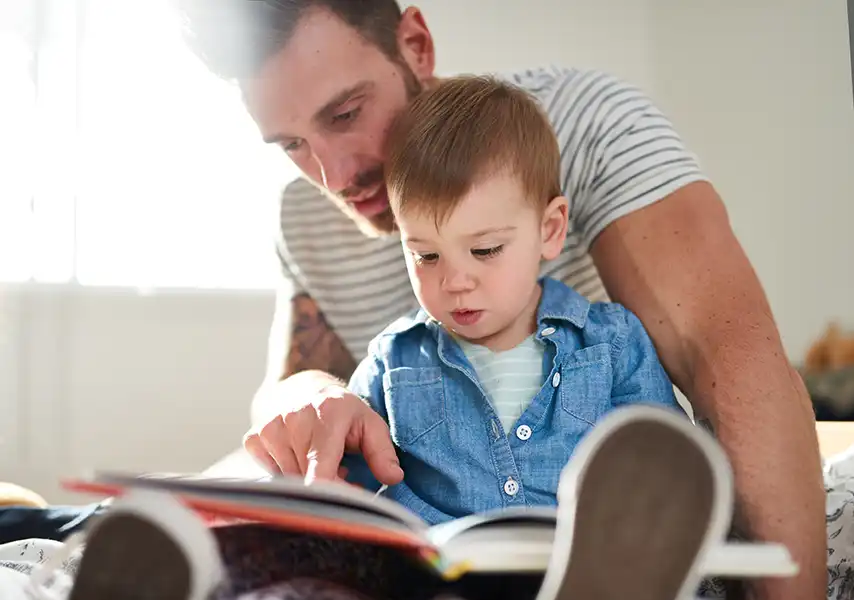 A young father reading a book to his toddler. They are sitting on his son's bedroom floor.